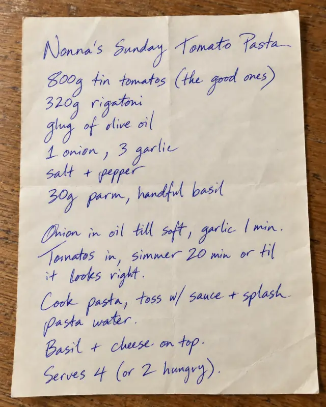 Nonna’s handwritten Sunday tomato pasta recipe in blue pen, photographed on a worn wooden kitchen table with the whole folded sheet visible.